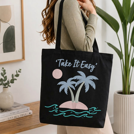 A woman wearing a beige ribbed sweater carries a black canvas tote bag with a tropical design. The bag features the text 'Take It Easy' in white script above two light blue palm trees on a pink island with turquoise waves.