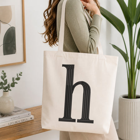 A woman wearing a sage green ribbed top carries a large off-white canvas tote bag with a bold black serif letter 'h' printed on the front, set in a bright, modern room with a wooden table, framed art, and a potted plant.
