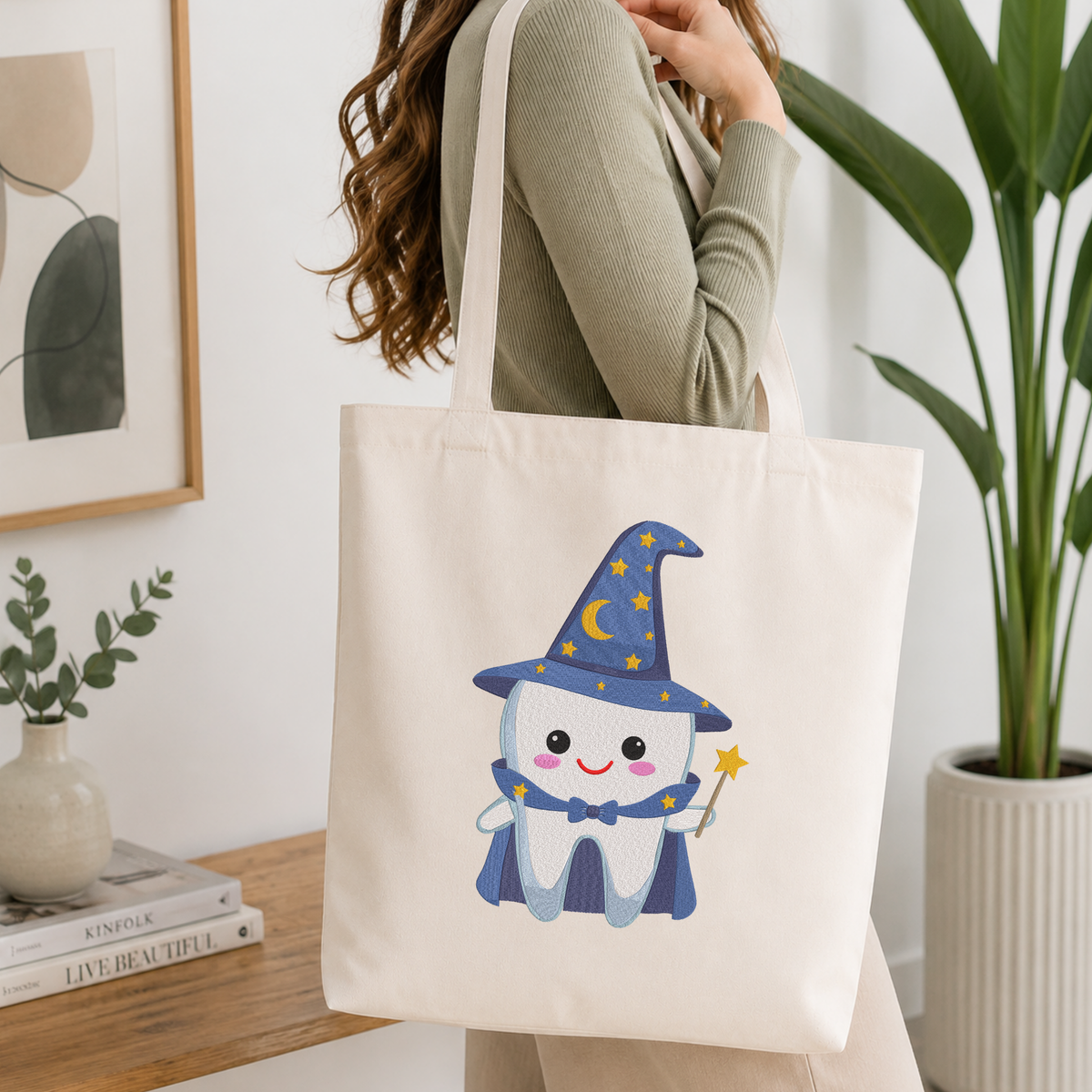 Woman carrying a white natural canvas tote bag featuring a cute wizard tooth machine embroidery design in a bright, modern interior setting