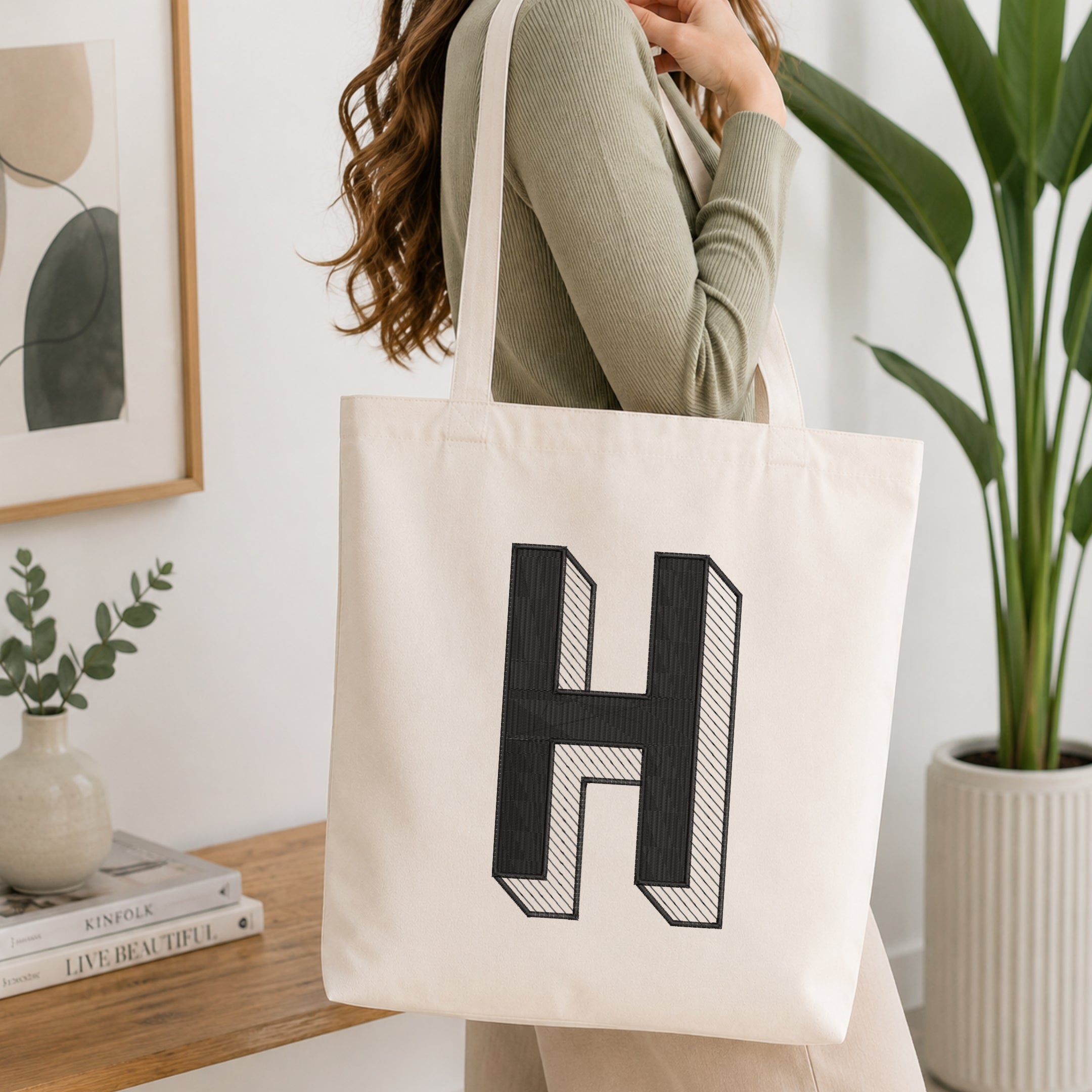 A woman wearing a sage green ribbed long-sleeve top carries a large off-white canvas tote bag with a bold black 3D-effect letter 'H' printed on the front. The scene is set in a bright, modern room with a wooden table, books, a framed print, and a potted green plant.
