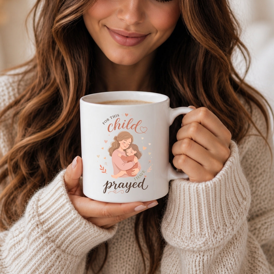 A hand holding a white ceramic mug featuring a mother-and-child illustration and the text "For this child I have prayed."