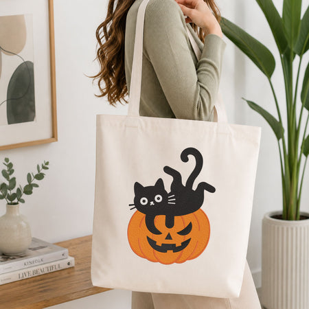 Woman carrying a white natural canvas tote bag featuring a cute black cat lying on a jack-o-lantern pumpkin machine embroidery design in a bright, modern interior setting