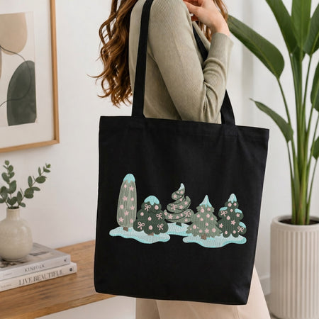 A woman wearing a beige ribbed sweater carries a black canvas tote bag with a whimsical embroidered winter tree design in mint green, teal, and white. The scene is set in a bright, modern room with a wooden table, books, a framed print, and a potted plant.