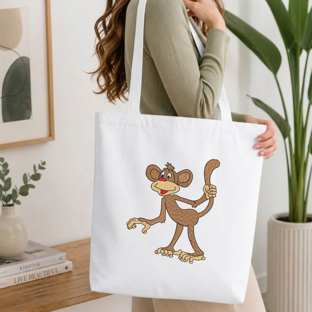 A woman wearing a sage green ribbed long-sleeve top carries a white canvas tote bag with a cartoon brown monkey graphic on the front, set in a bright, modern room with a wooden table, books, and a potted plant.