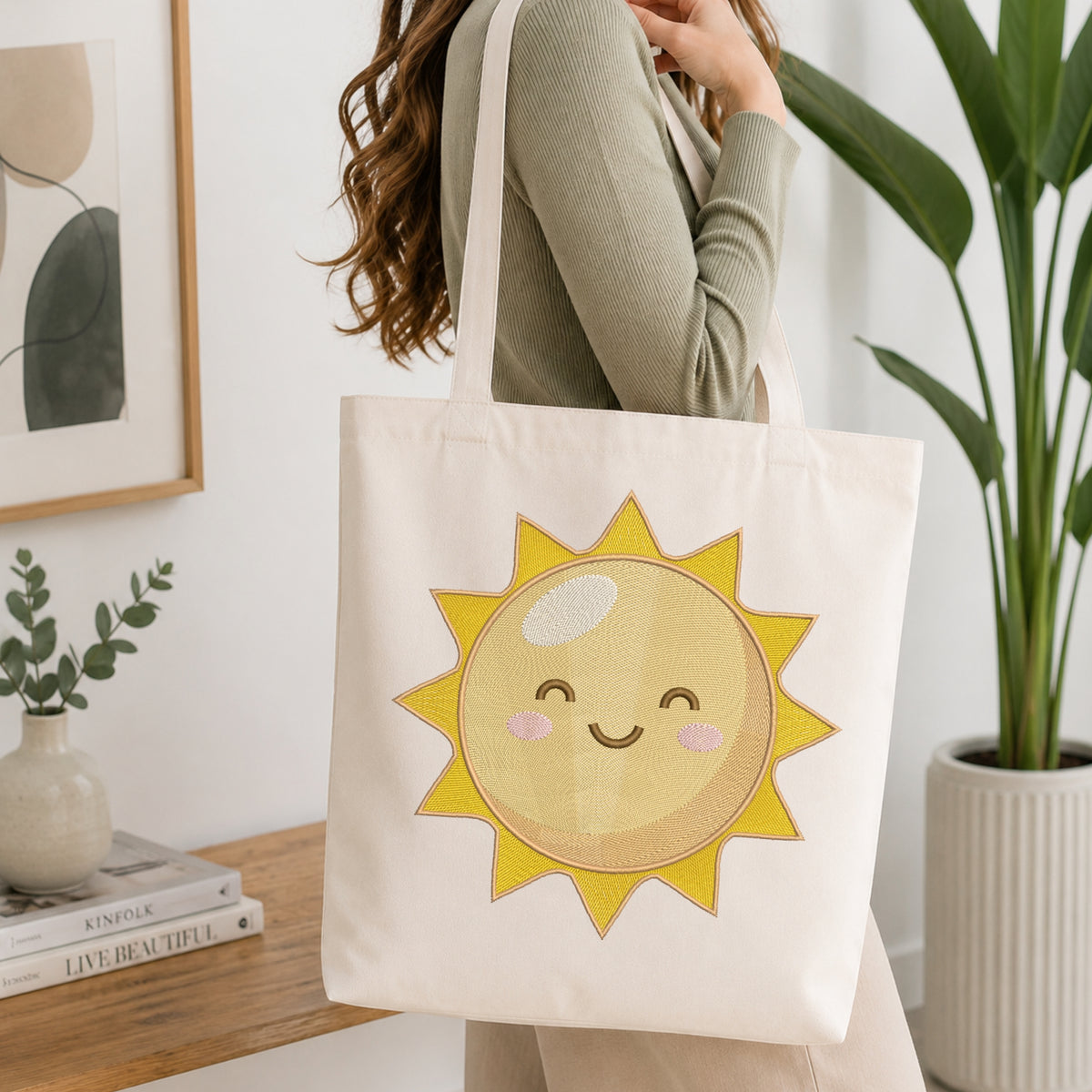 A woman wearing a sage green ribbed long-sleeve top carries a cream canvas tote bag with a large embroidered smiling sun design on the front, set in a bright, modern room with a wooden table, books, and a potted plant.
