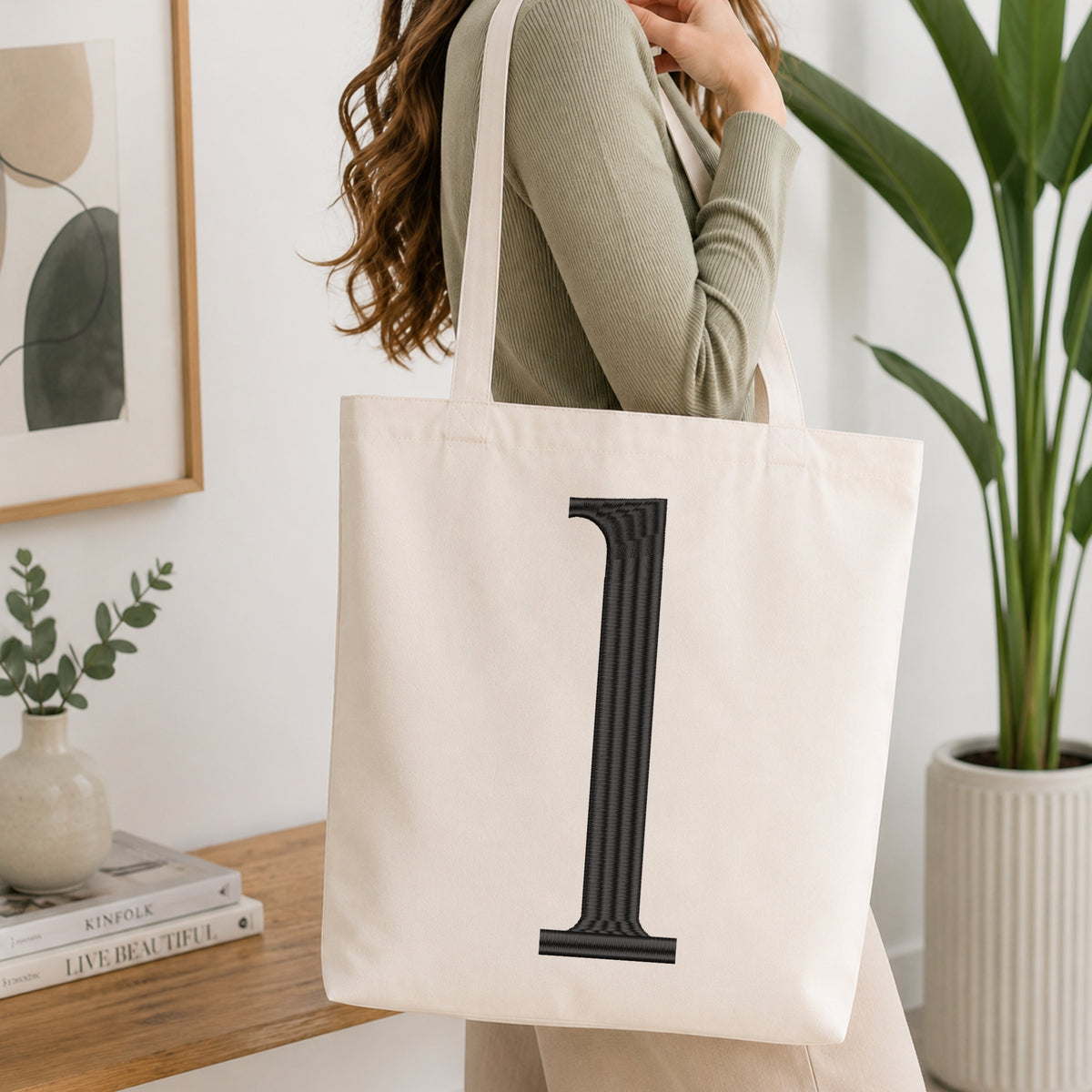 A woman wearing a sage green ribbed long-sleeve top carries a cream canvas tote bag with a large black serif letter 'L' printed on the front, set in a bright, modern room with a wooden console table, framed art, and a potted plant.
