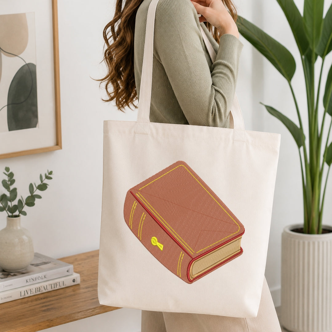 A woman wearing a sage green ribbed sweater carries a cream canvas tote bag with a graphic of a brown leather-bound book with gold trim. The background is a bright, modern room with a wooden table, books, a small plant, and a large green potted plant.