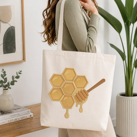 A woman wearing a sage green ribbed sweater carries a cream canvas tote bag with a printed honeycomb and honey dipper design in gold and brown, set against a bright, modern interior background with a wooden table, framed art, and a potted plant.