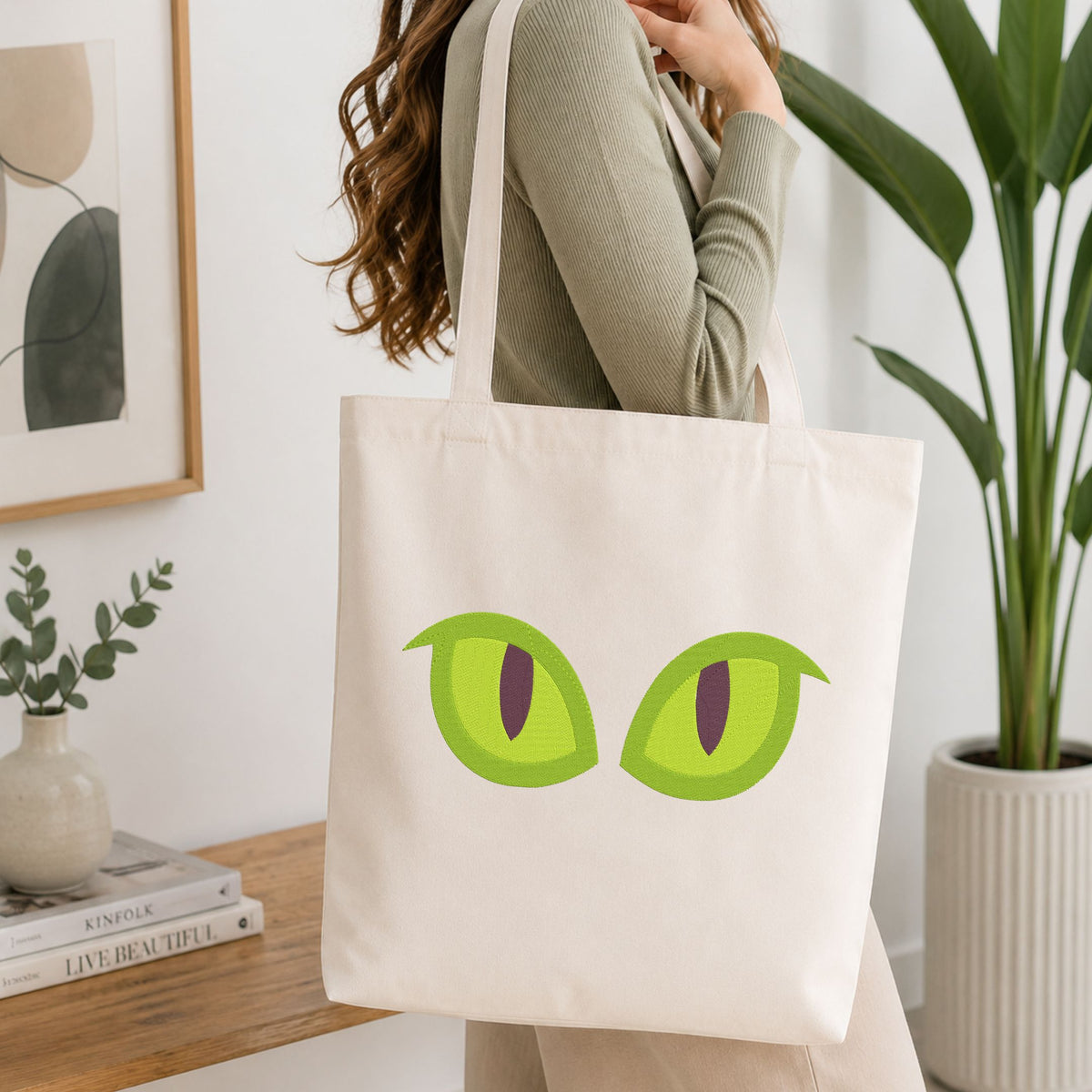 A woman wearing a sage green ribbed top carries a cream canvas tote bag featuring a graphic of two green cartoon eyes with dark purple pupils, set against a bright, modern room background with a wooden table and potted plants.