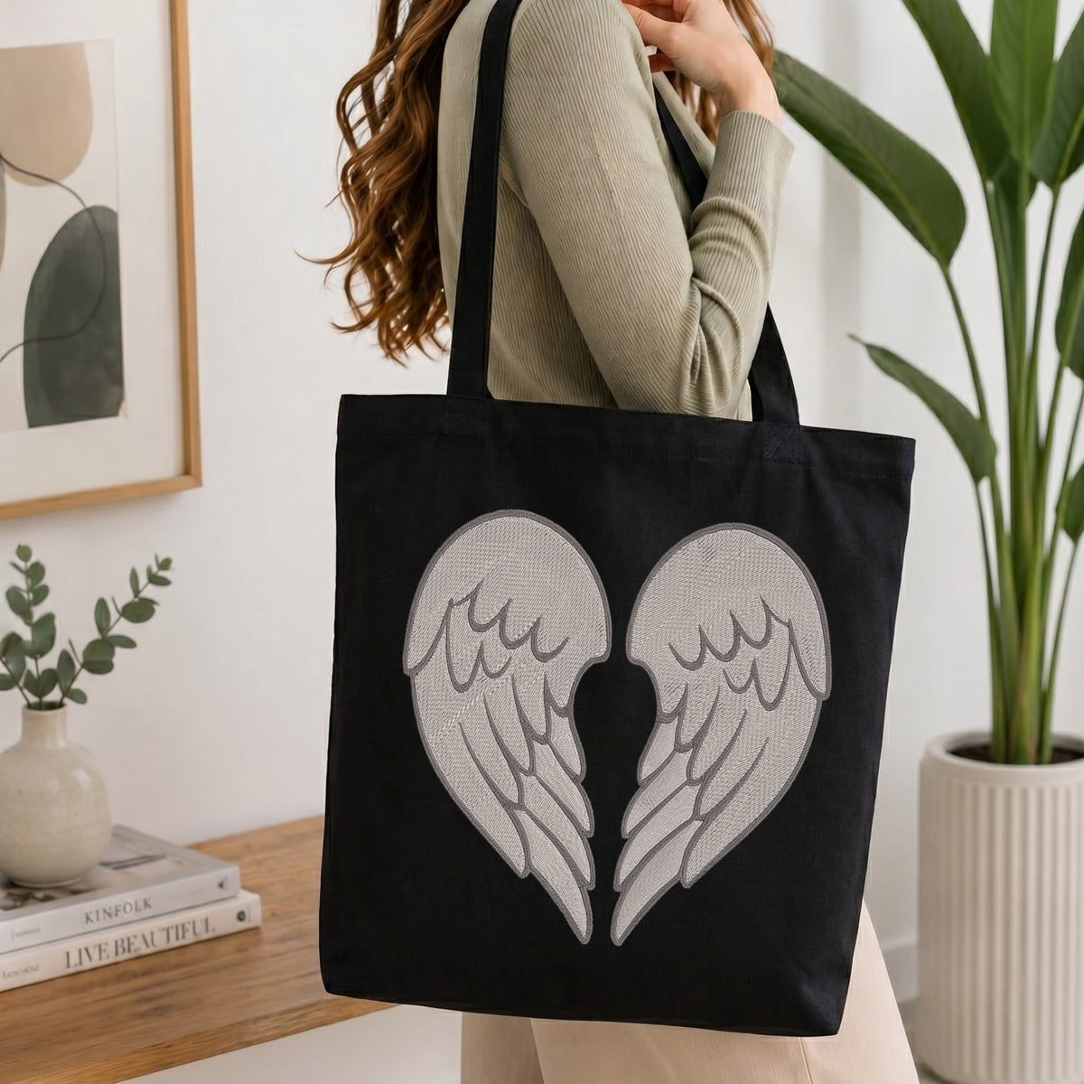 A woman wearing a beige ribbed sweater carries a black canvas tote bag with a large gray embroidered broken-heart wings design on the front, set in a bright, modern room with a wooden table, books, and a potted plant.