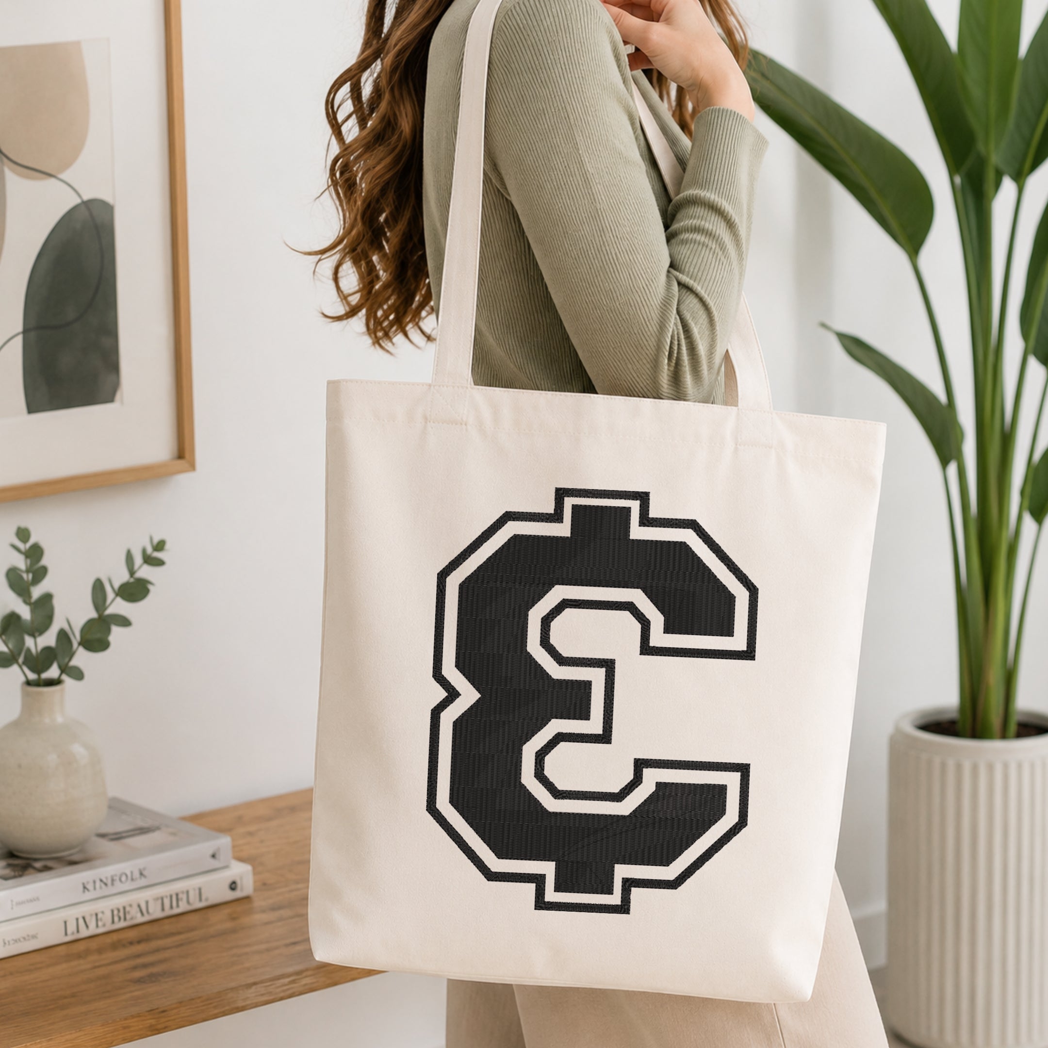 A woman wearing a sage green ribbed long-sleeve top carries a cream canvas tote bag with a large black and white pixelated letter C on the front, set in a bright, modern room with a wooden console table and a potted plant.