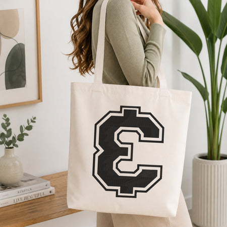 A woman wearing a sage green ribbed long-sleeve top carries a cream canvas tote bag with a large black and white pixelated letter C on the front, set in a bright, modern room with a wooden console table and a potted plant.