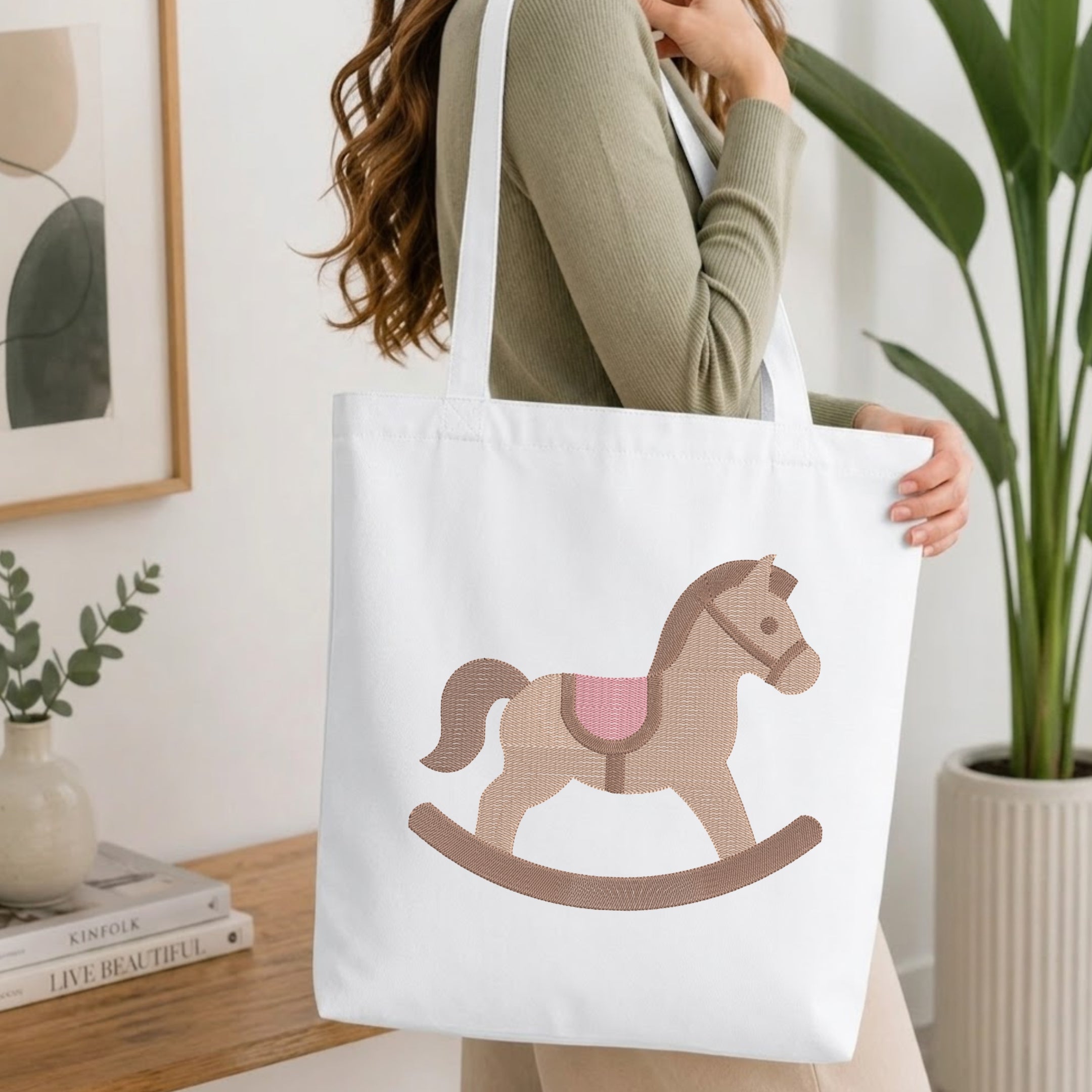 A woman wearing a sage green ribbed long-sleeve top carries a white canvas tote bag featuring a retro-style rocking horse illustration in tan, brown, and pink. The setting is a bright, modern room with a wooden table, books, a framed print, and a potted green plant.