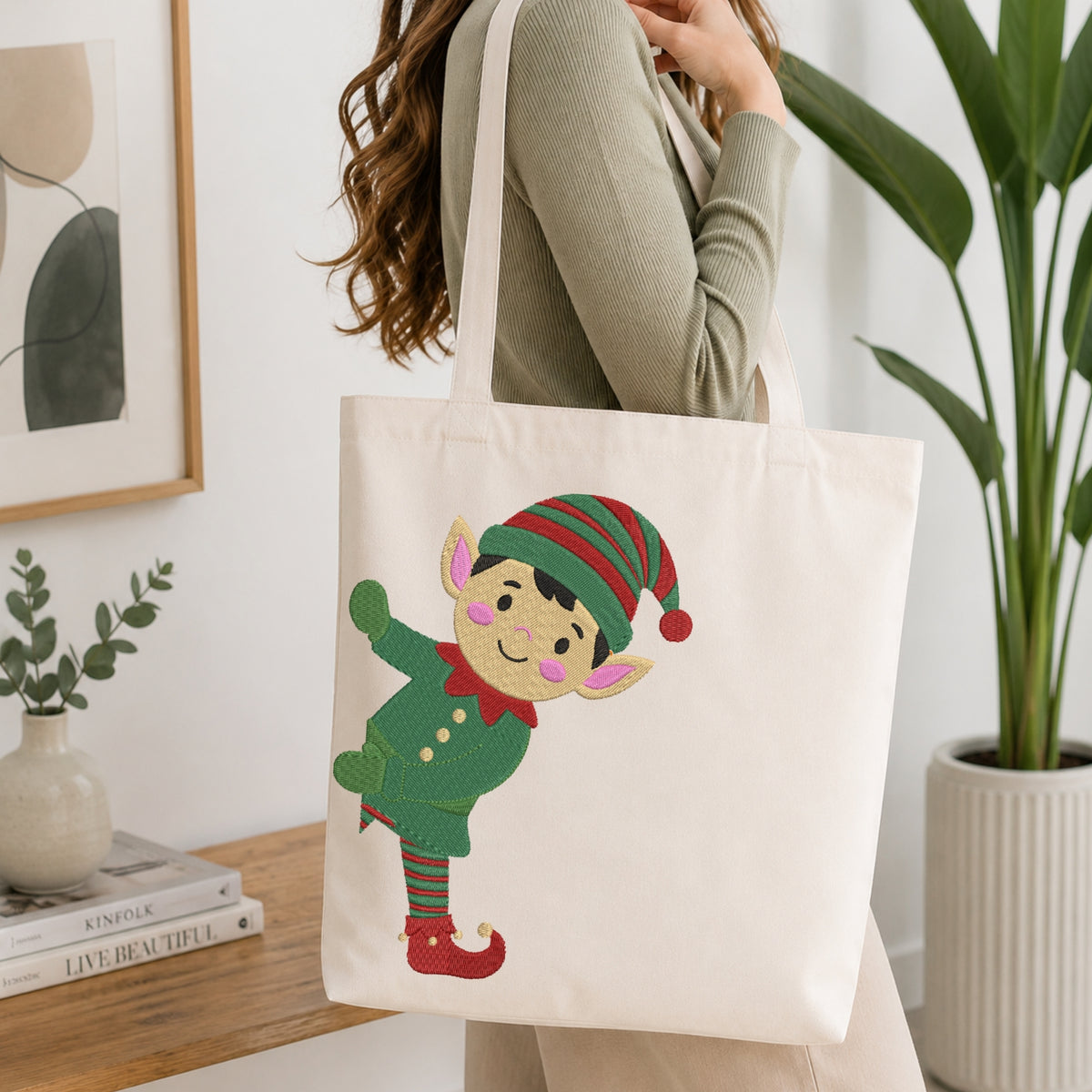 A woman carries a cream canvas tote bag with a colorful embroidered elf character on the front, set in a bright, modern room with a wooden table and potted plants.