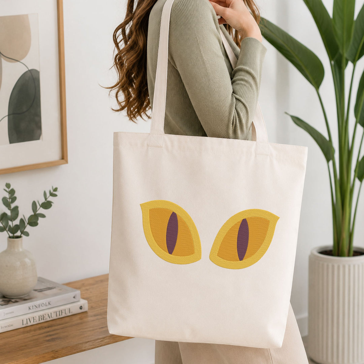 A woman wearing a sage green ribbed long-sleeve top carries a cream canvas tote bag featuring a graphic of two yellow cat eyes with purple pupils, set in a bright, modern room with a wooden table and potted plants.