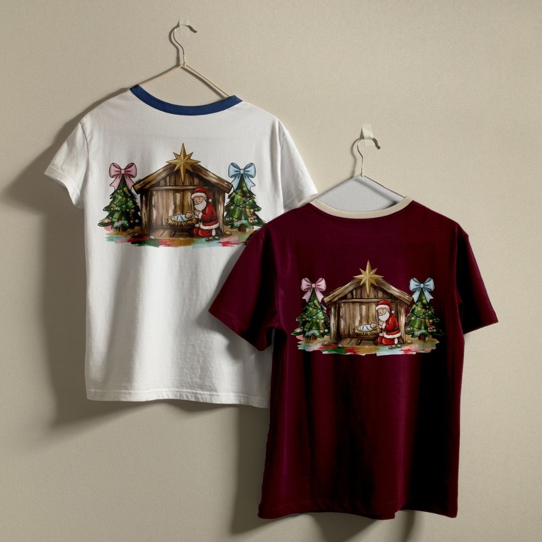 Two Christmas-themed t-shirts hanging on hangers against a plain beige wall. The left shirt is white with a blue collar and shows the back graphic of a nativity scene with Santa.