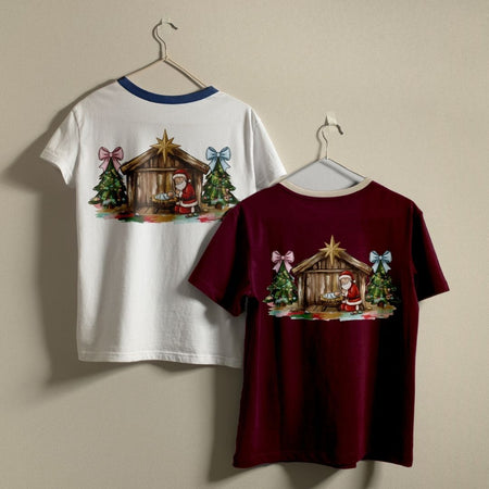 Two Christmas-themed t-shirts hanging on hangers against a plain beige wall. The left shirt is white with a blue collar and shows the back graphic of a nativity scene with Santa.