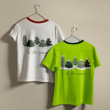 Two Christmas-themed ringer t-shirts hanging on metal hangers against a plain beige wall. The left shirt is white with a red collar trim and shows a back graphic of decorated trees in a snowy landscape with the text 'it's the season'.