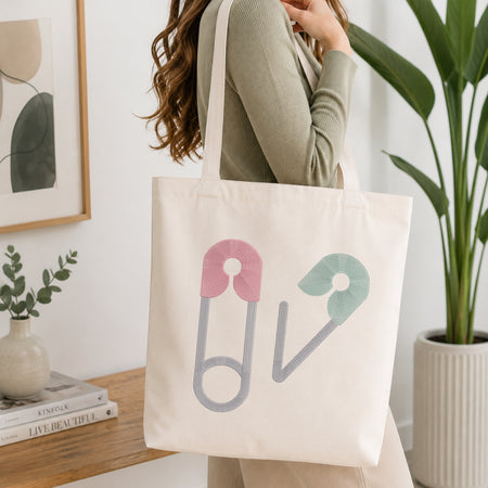 A woman wearing a sage green ribbed long-sleeve top carries a cream canvas tote bag with a graphic of two safety pins in pink and mint green on a gray outline, set in a bright, modern room with a wooden table and potted plants.