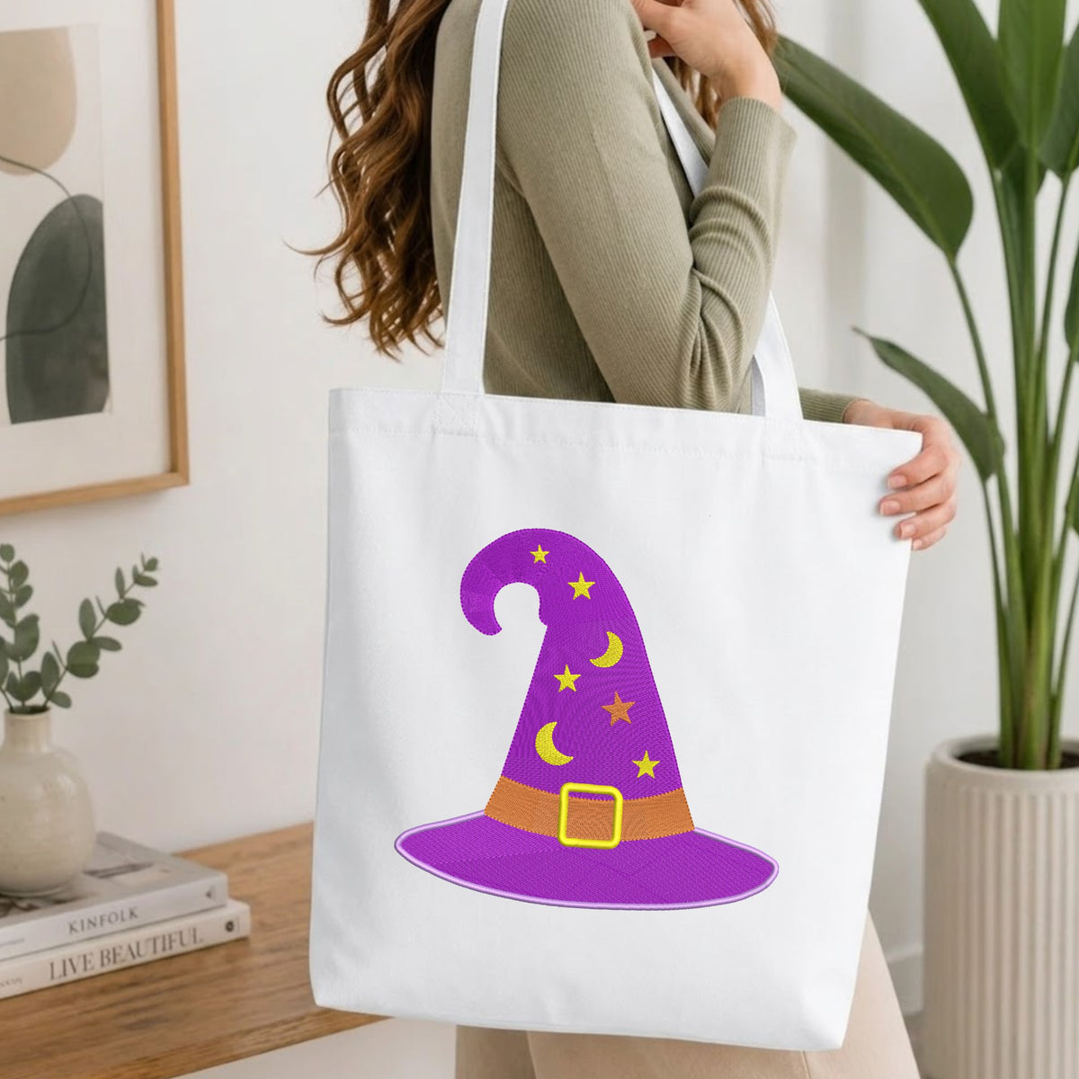 Woman carrying a white natural canvas tote bag featuring an embroidered purple witch hat design in a bright, modern interior setting.