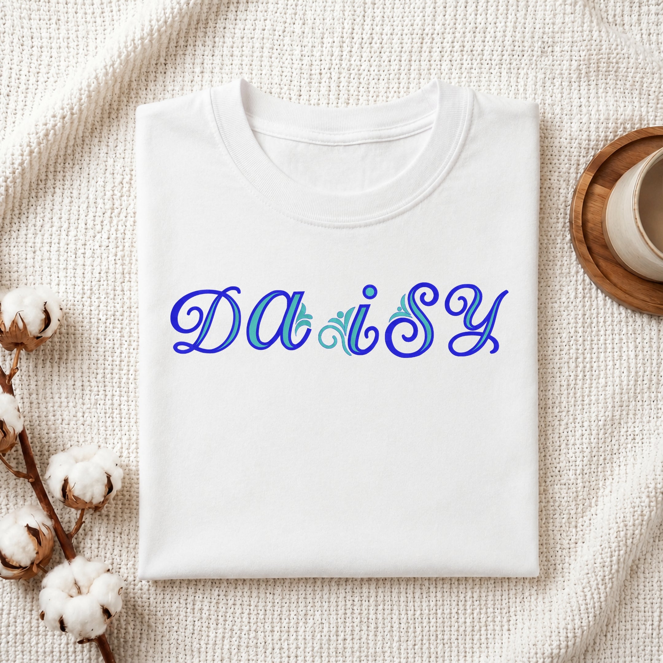 A folded white crewneck t-shirt with the word 'DAISY' printed across the chest in blue cursive script with light blue floral accents, styled on a textured cream fabric background with cotton bolls and a coffee cup.