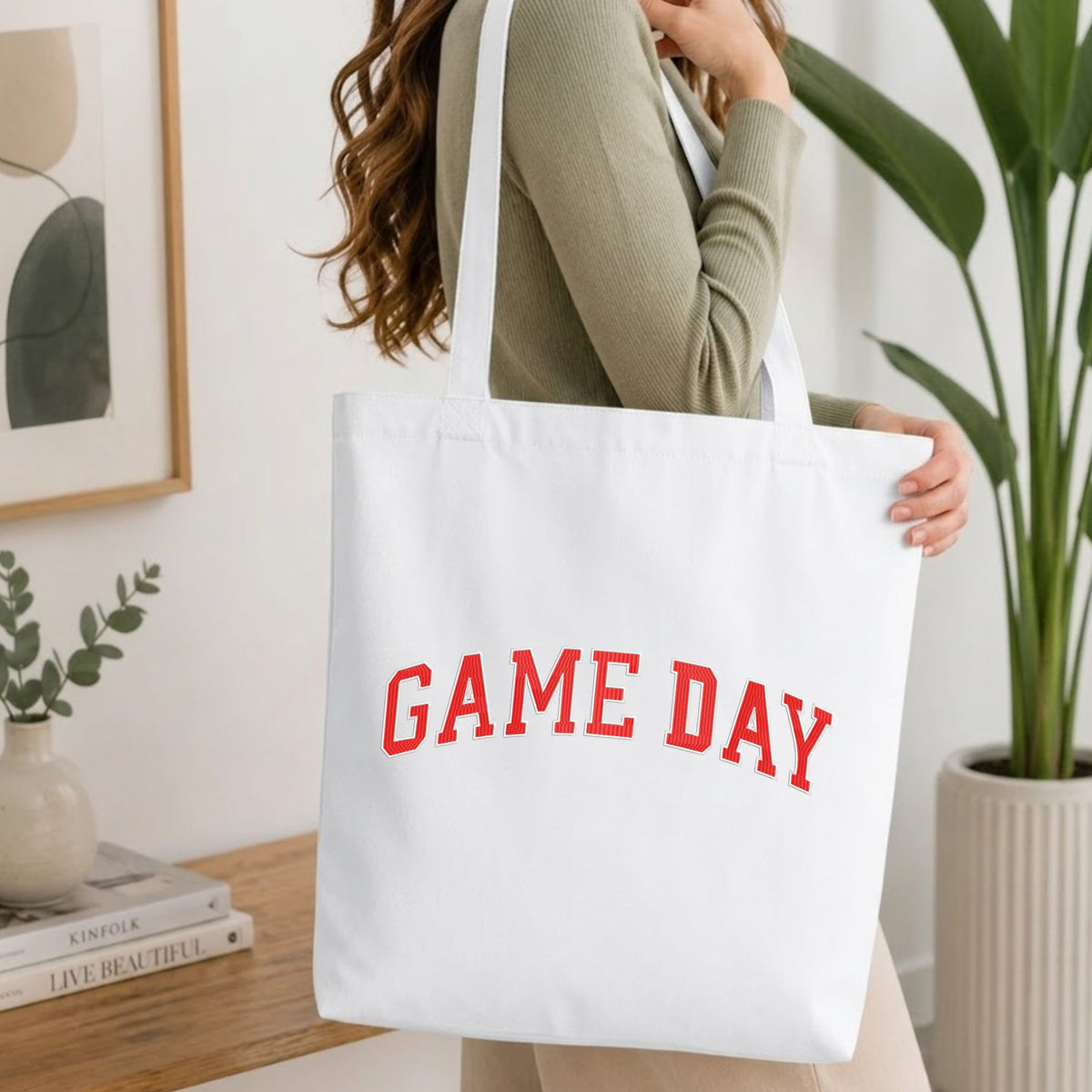 Woman carrying a white natural canvas tote bag featuring an embroidered red "GAME DAY" collegiate block text design in a bright, modern interior setting.