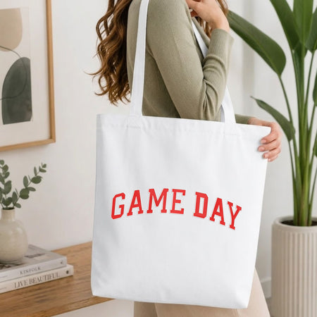 Woman carrying a white natural canvas tote bag featuring an embroidered red "GAME DAY" collegiate block text design in a bright, modern interior setting.