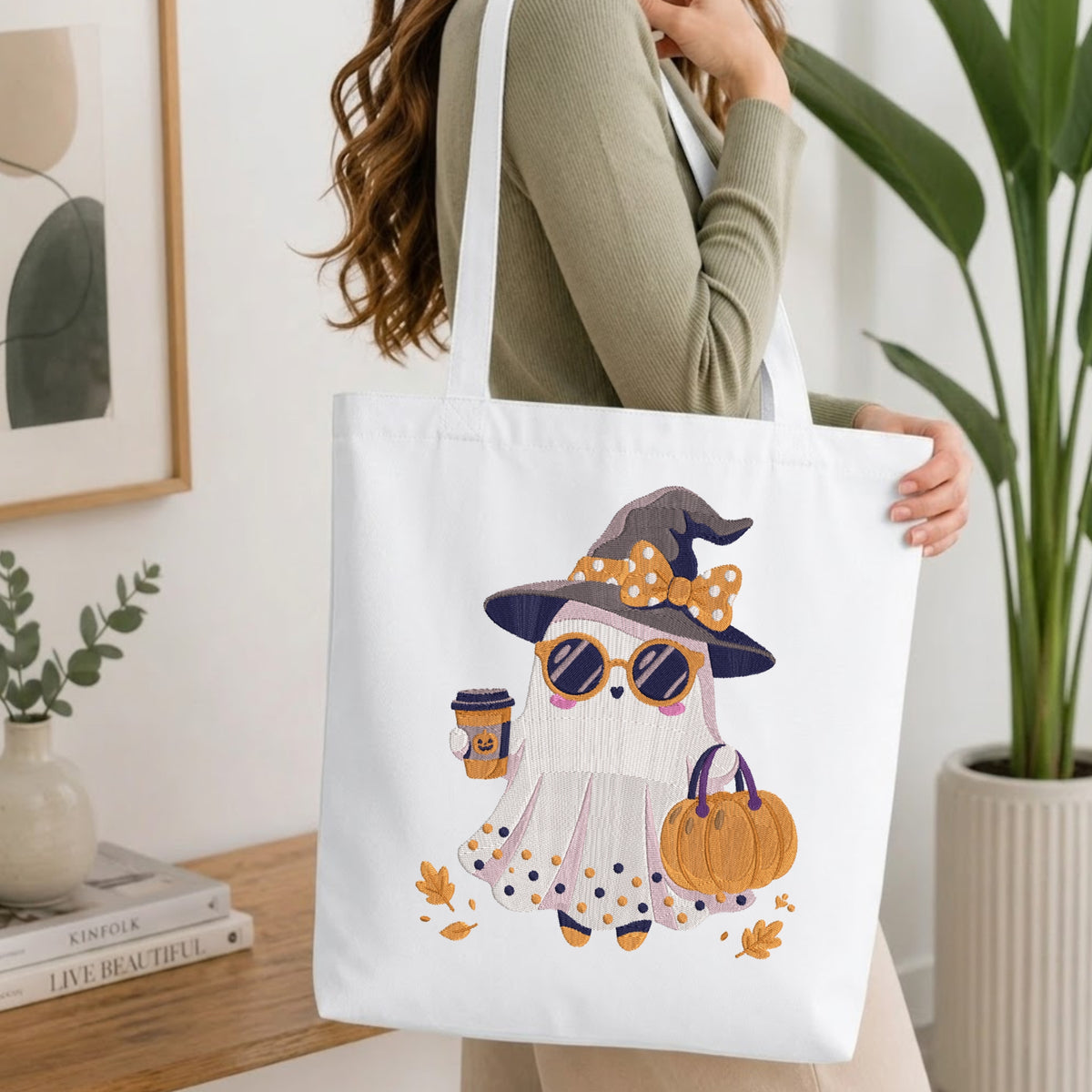 A woman holds a white canvas tote bag featuring a cute cartoon ghost dressed as a witch with a purple hat, orange polka-dot bow, sunglasses, a coffee cup, and a pumpkin. The scene is set in a bright, modern room with a wooden table, books, and a potted plant.