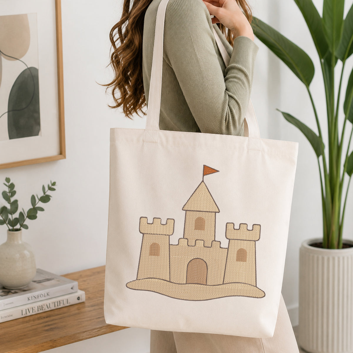 A woman wearing a sage green ribbed sweater carries a cream canvas tote bag with a tan and brown castle graphic printed on the front, set in a bright, modern room with a wooden table, books, and a potted plant.
