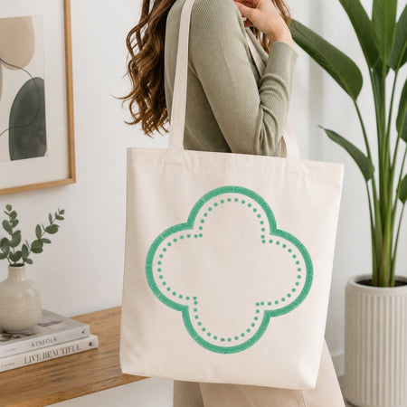 A woman wearing a sage green ribbed sweater carries a cream canvas tote bag with a mint green quatrefoil outline graphic, set in a bright, modern room with a wooden table, books, and a potted plant.
