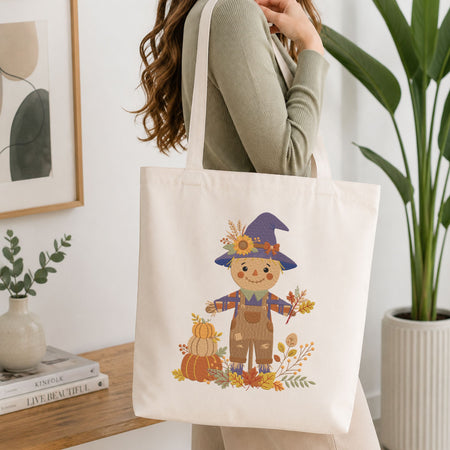 Woman carrying a white natural canvas tote bag featuring a cute autumn scarecrow machine embroidery design in a bright, modern interior setting