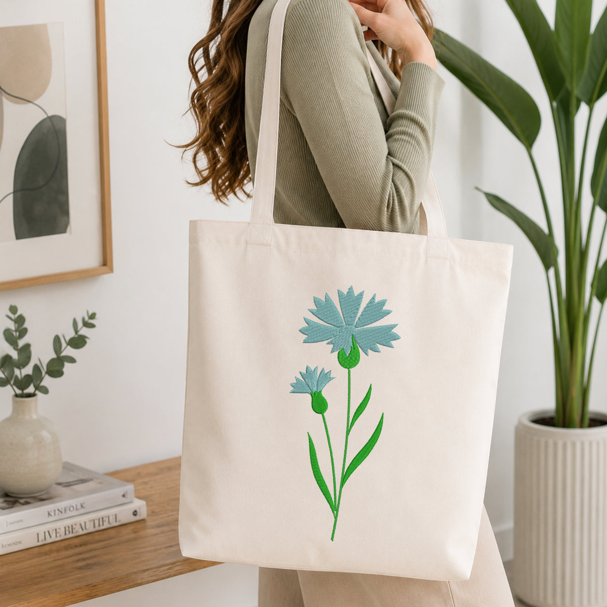 A woman wearing a sage green ribbed sweater carries a cream canvas tote bag with a light blue and green floral print, set in a bright, modern room with a wooden table and potted plants.