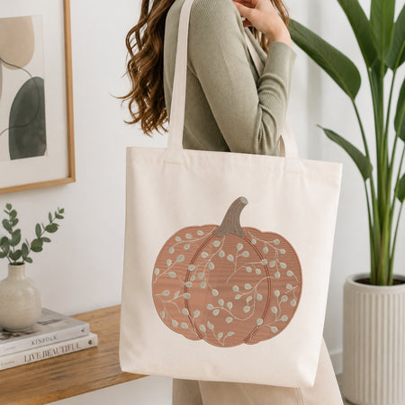 A woman wearing a sage green ribbed sweater carries a cream canvas tote bag featuring a large embroidered peach pumpkin with pale green leaves. The scene is set in a bright, modern room with a wooden table, books, a framed print, and a potted plant.
