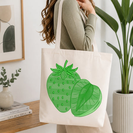 A person wearing a sage green ribbed sweater carries a cream canvas tote bag with a green strawberry illustration, set in a bright, modern room with a wooden table, books, and a potted plant.