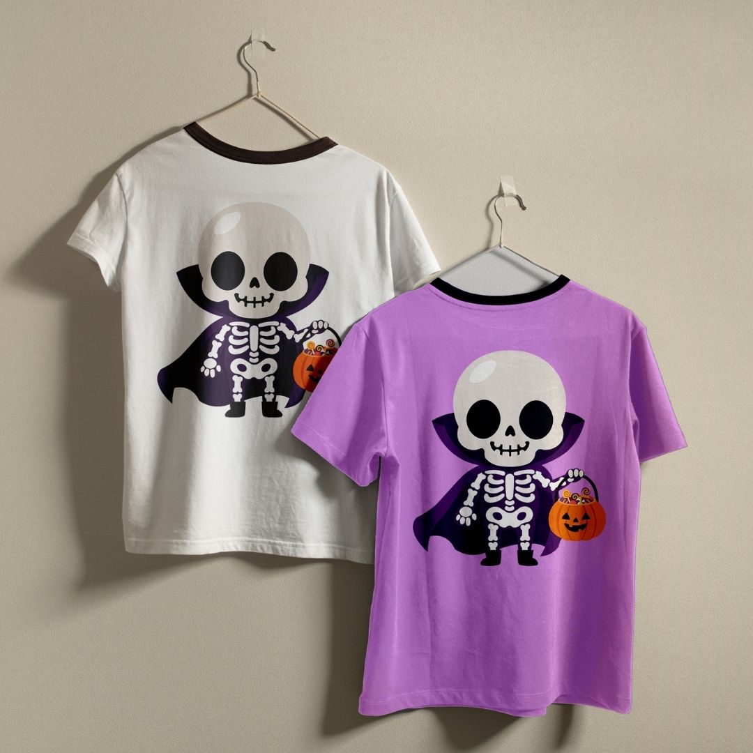 Two children's ringer t-shirts with a Halloween vampire skeleton graphic, shown on hangers against a plain beige wall. One shirt is white with a dark brown collar, and the other is purple with a black collar.