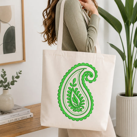 A person wearing a ribbed sage green long-sleeve top carries a large off-white canvas tote bag with a prominent green paisley pattern. The setting is a bright, modern room with a wooden console table, a framed print, a small vase, and a potted plant.