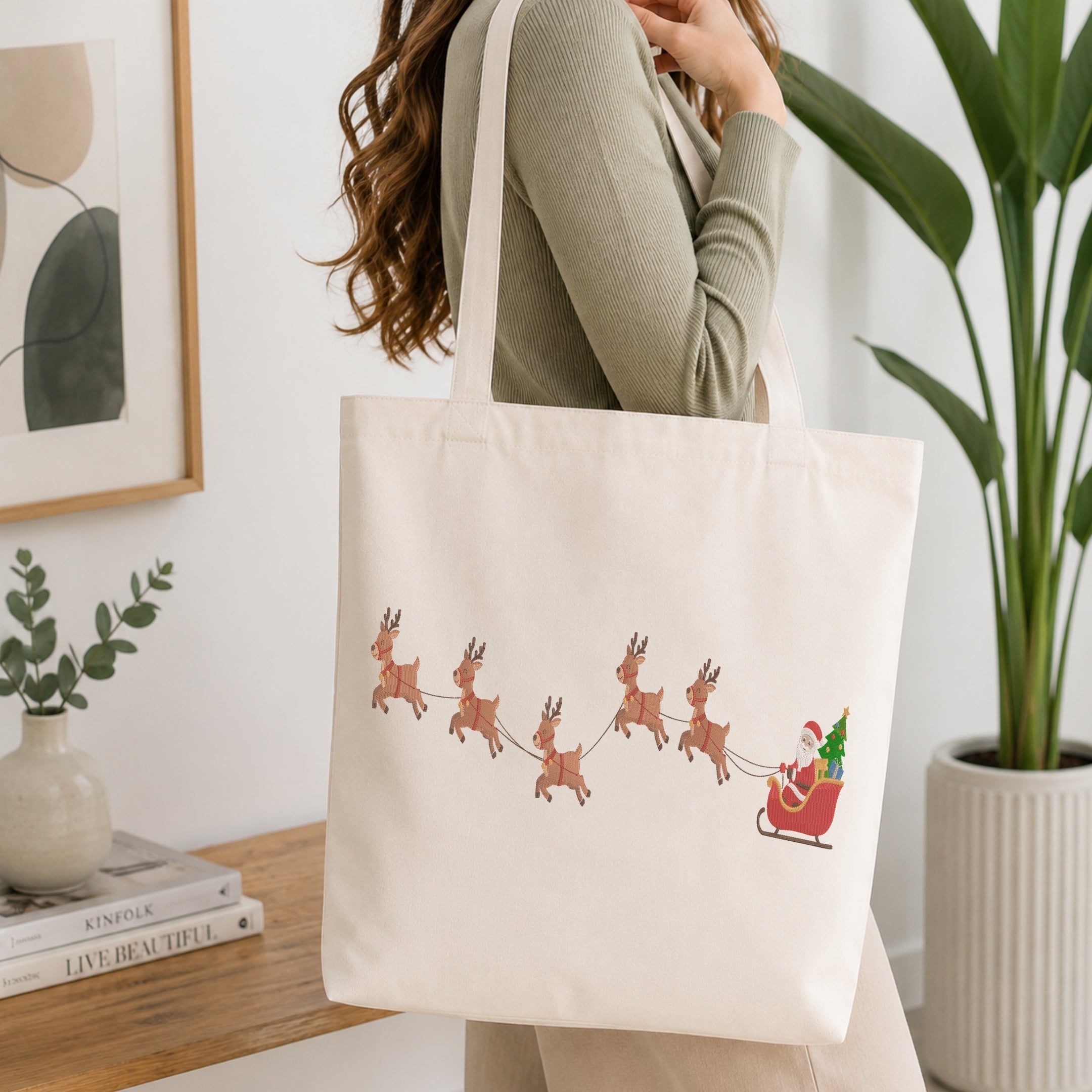 A woman wearing a sage green ribbed sweater carries a cream canvas tote bag with a printed illustration of Santa Claus in a red sleigh pulled by brown reindeer, set against a bright, modern interior background with a potted plant and books.