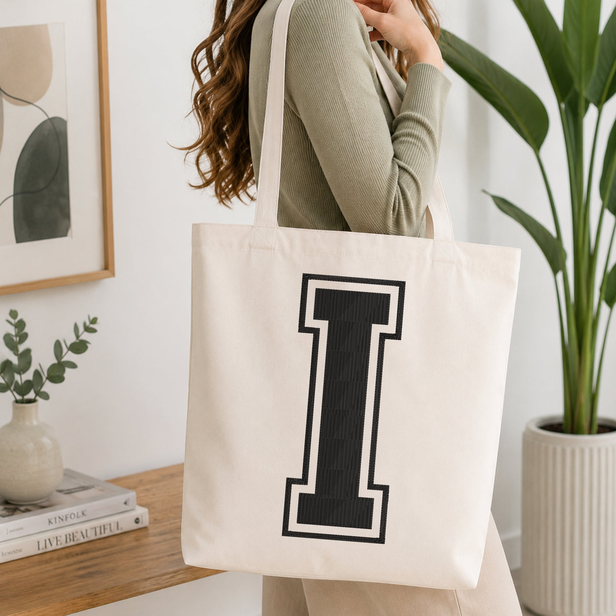 A woman wearing a sage green ribbed long-sleeve top carries a large off-white canvas tote bag with a bold black block letter 'I' outlined in white on the front, set in a bright, modern room with a wooden console table, framed art, and a potted plant.
