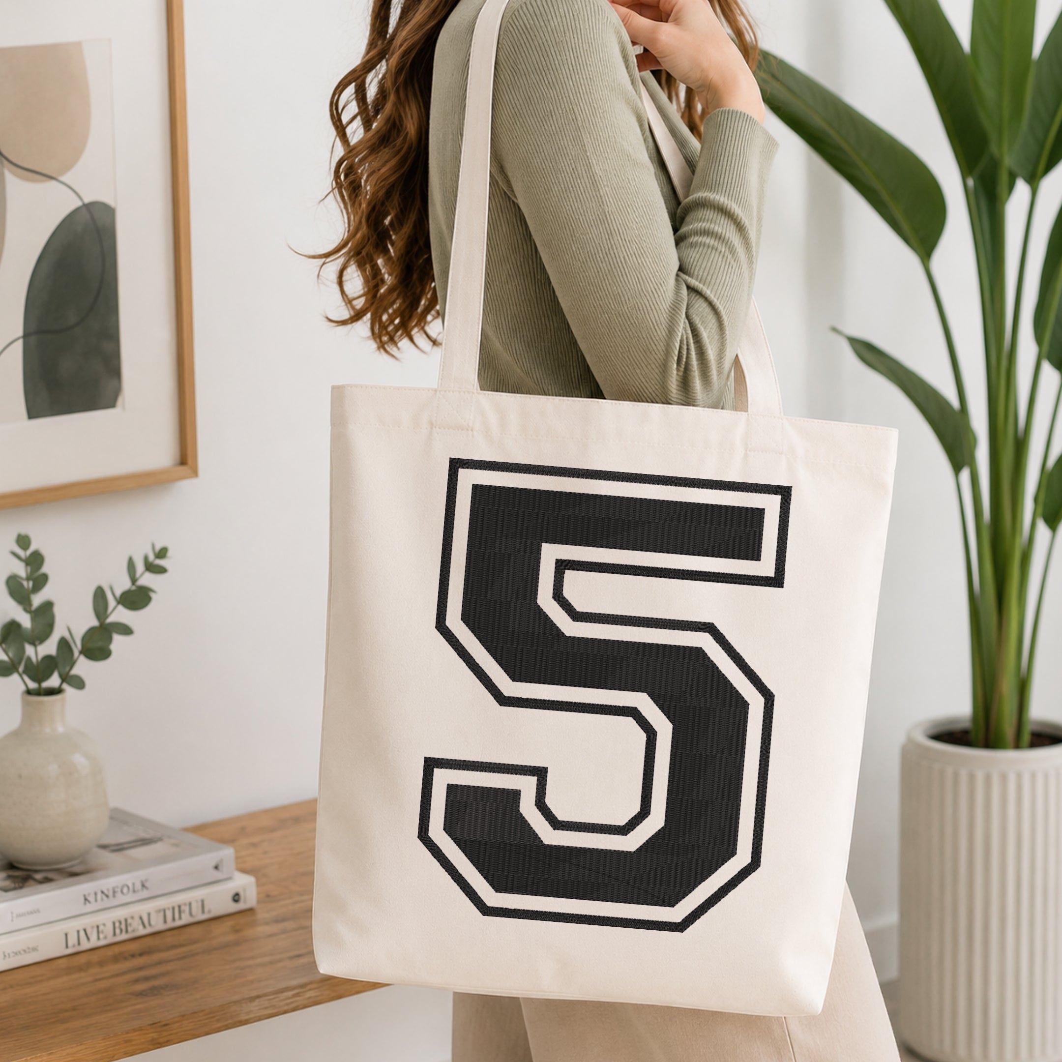 A person wearing a sage green ribbed long-sleeve top carries a cream canvas tote bag with a large black and white number 5 printed on the front, set in a bright, modern room with a wooden console table and a potted plant.