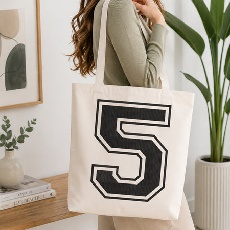 A person wearing a sage green ribbed long-sleeve top carries a cream canvas tote bag with a large black and white number 5 printed on the front, set in a bright, modern room with a wooden console table and a potted plant.