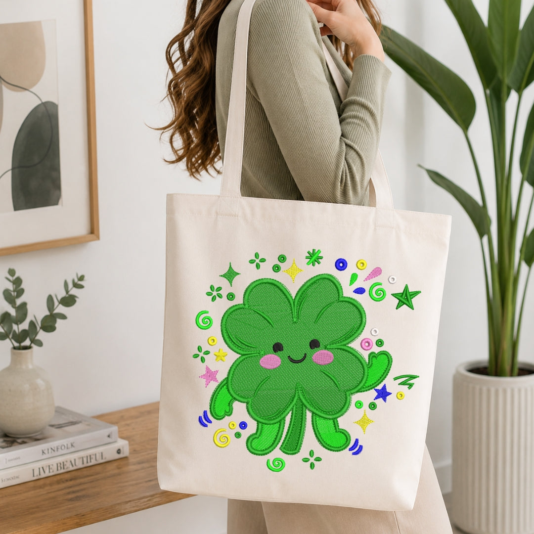 A person wearing a ribbed sage green long-sleeve top carries a cream canvas tote bag. The bag features a colorful embroidered design of a smiling green four-leaf clover surrounded by playful stars, swirls, and dots in green, yellow, pink, blue, and purple.