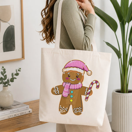 A woman wearing a sage green ribbed sweater carries a cream canvas tote bag with a colorful gingerbread man graphic. The scene is set in a bright, modern room with a wooden table, a potted plant, and a framed print.