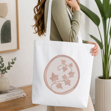 A woman wearing a sage green ribbed long-sleeve top carries a white canvas tote bag with a circular rose gold ivy pattern. The background is a bright, modern room with a wooden table, books, a framed print, and a potted plant.