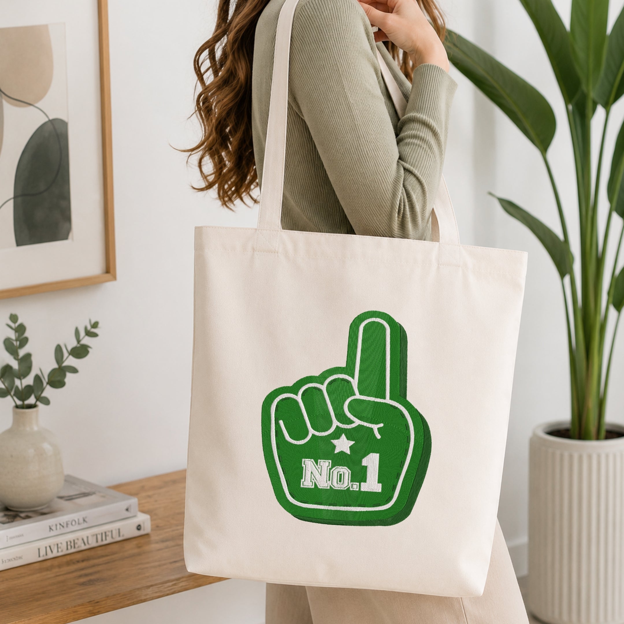 A woman wearing a sage green ribbed long-sleeve top carries a cream canvas tote bag with a green foam finger graphic that says 'No. 1' in white.