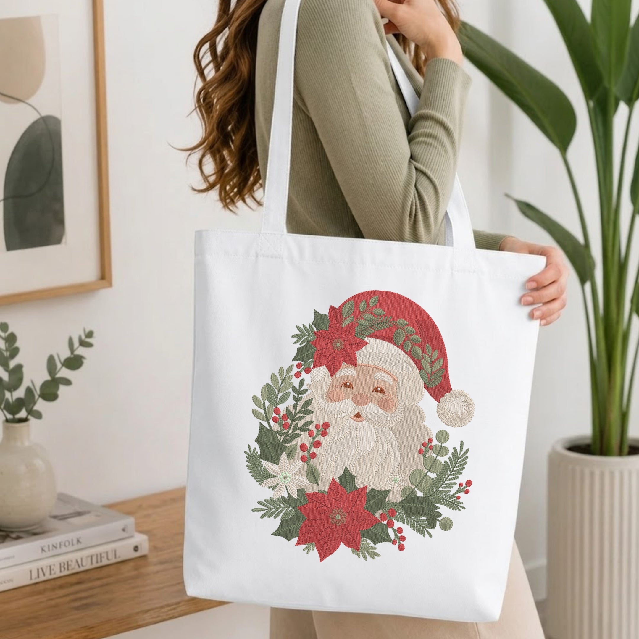 A woman wearing a sage green ribbed long-sleeve top carries a white canvas tote bag decorated with a large embroidered Christmas Santa design featuring a red hat, white beard, and green wreath.