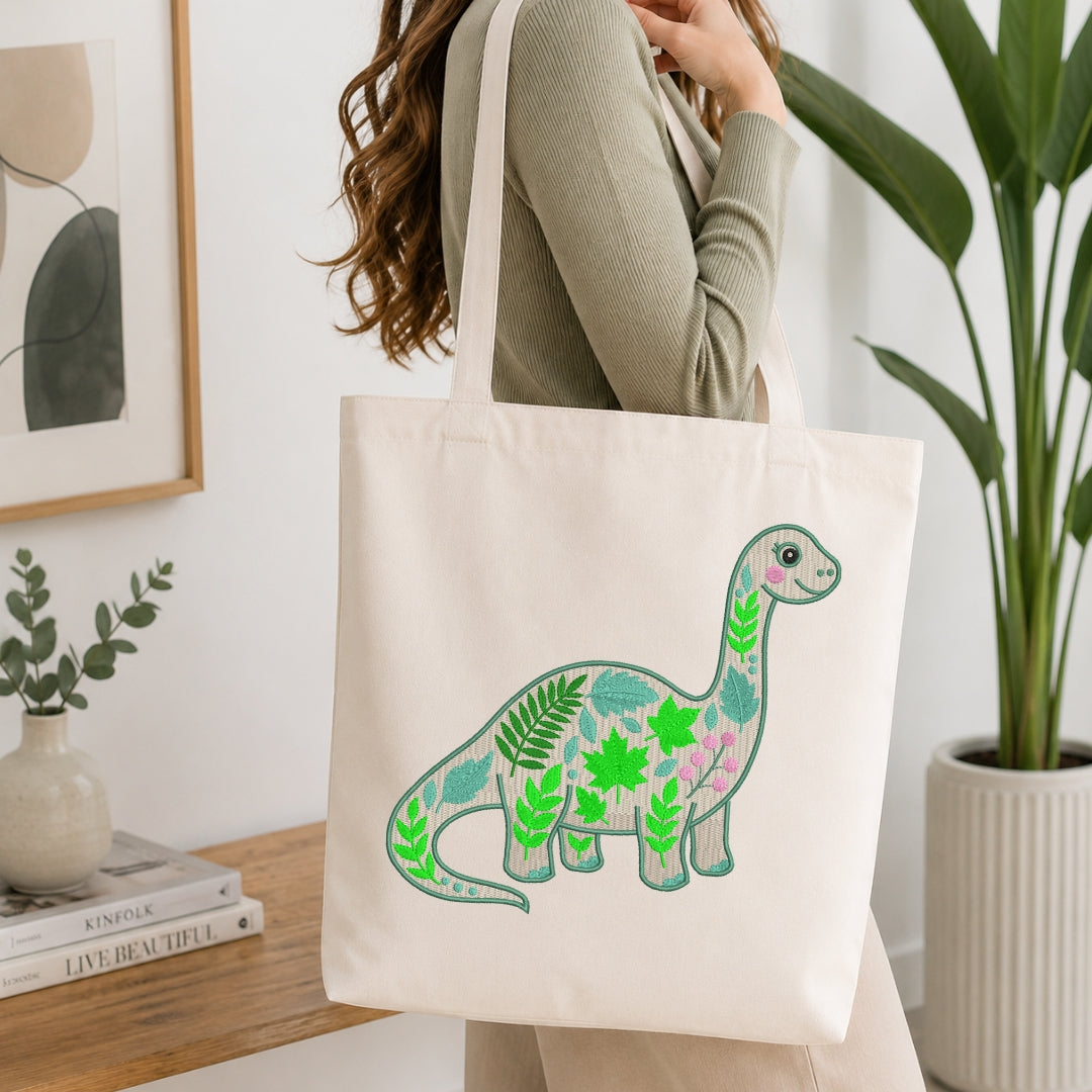 A woman wearing a sage green ribbed sweater carries a cream canvas tote bag with a colorful embroidered dinosaur design featuring green and pink leaves.