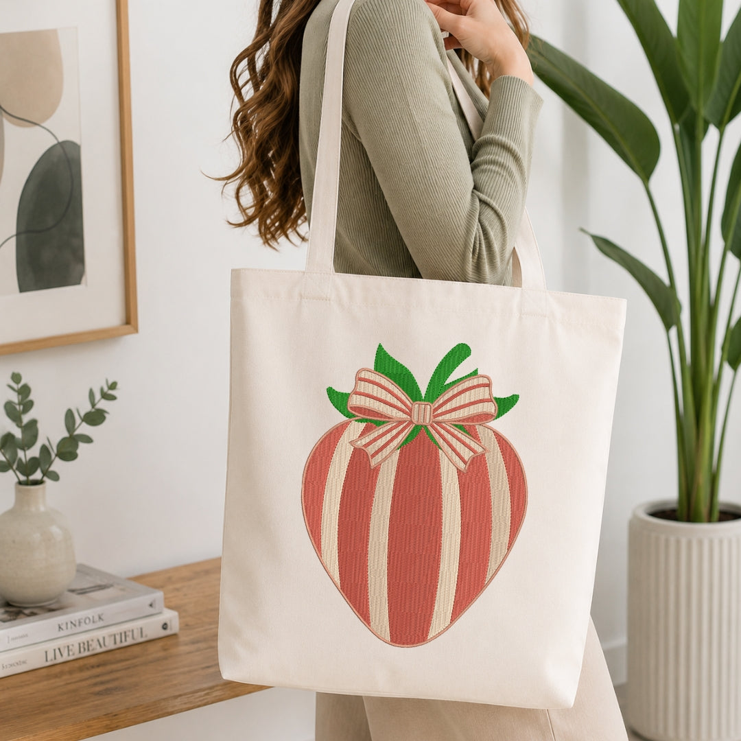 A woman wearing a sage green ribbed sweater carries a cream canvas tote bag with a large embroidered strawberry design featuring red and cream vertical stripes and a pink bow with green leaves.