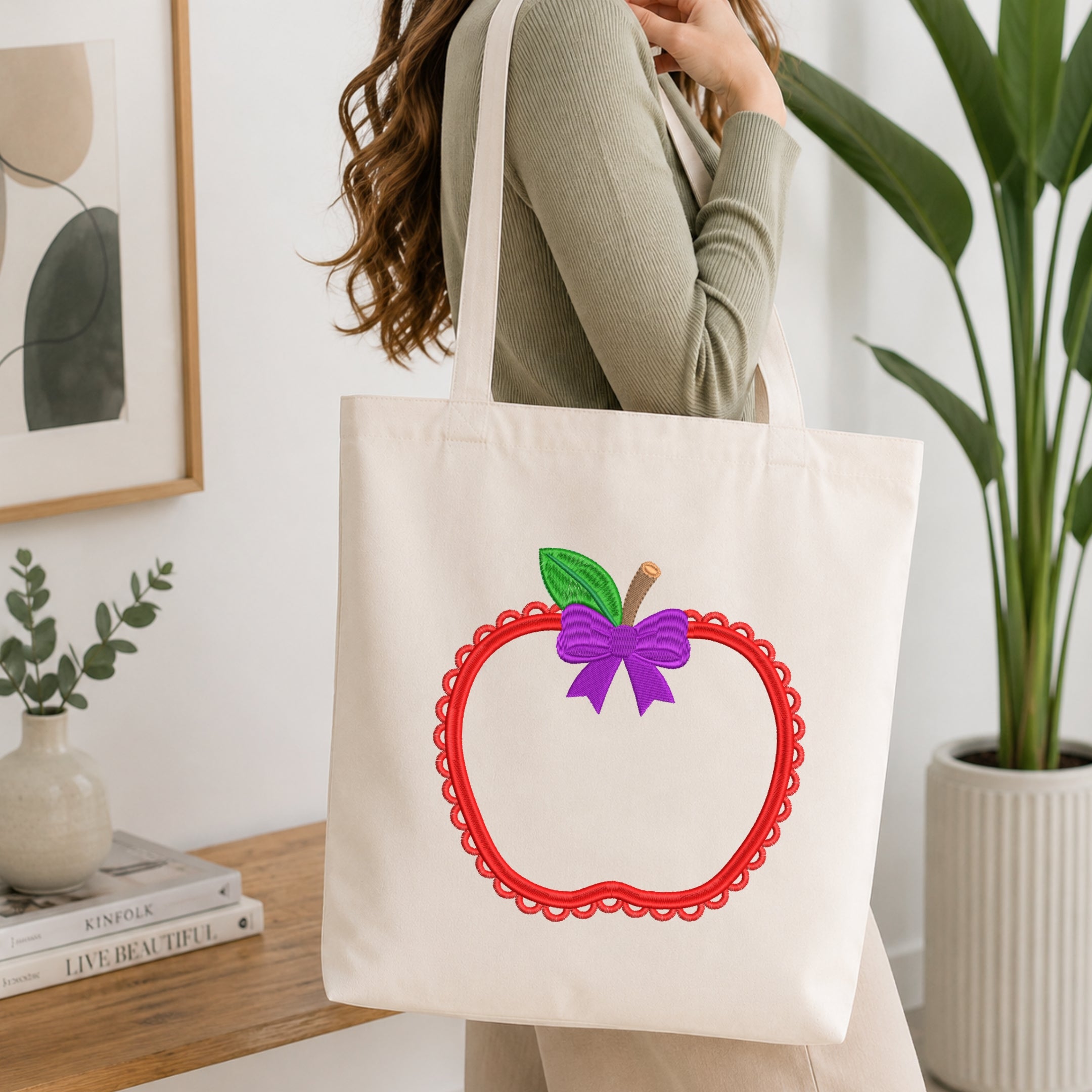 A woman wearing a sage green ribbed long-sleeve top carries a cream canvas tote bag with a large applique design of a red scalloped apple outline, a green leaf, and a purple bow. The scene is set in a bright, modern room with a wooden table, books, and a potted plant.