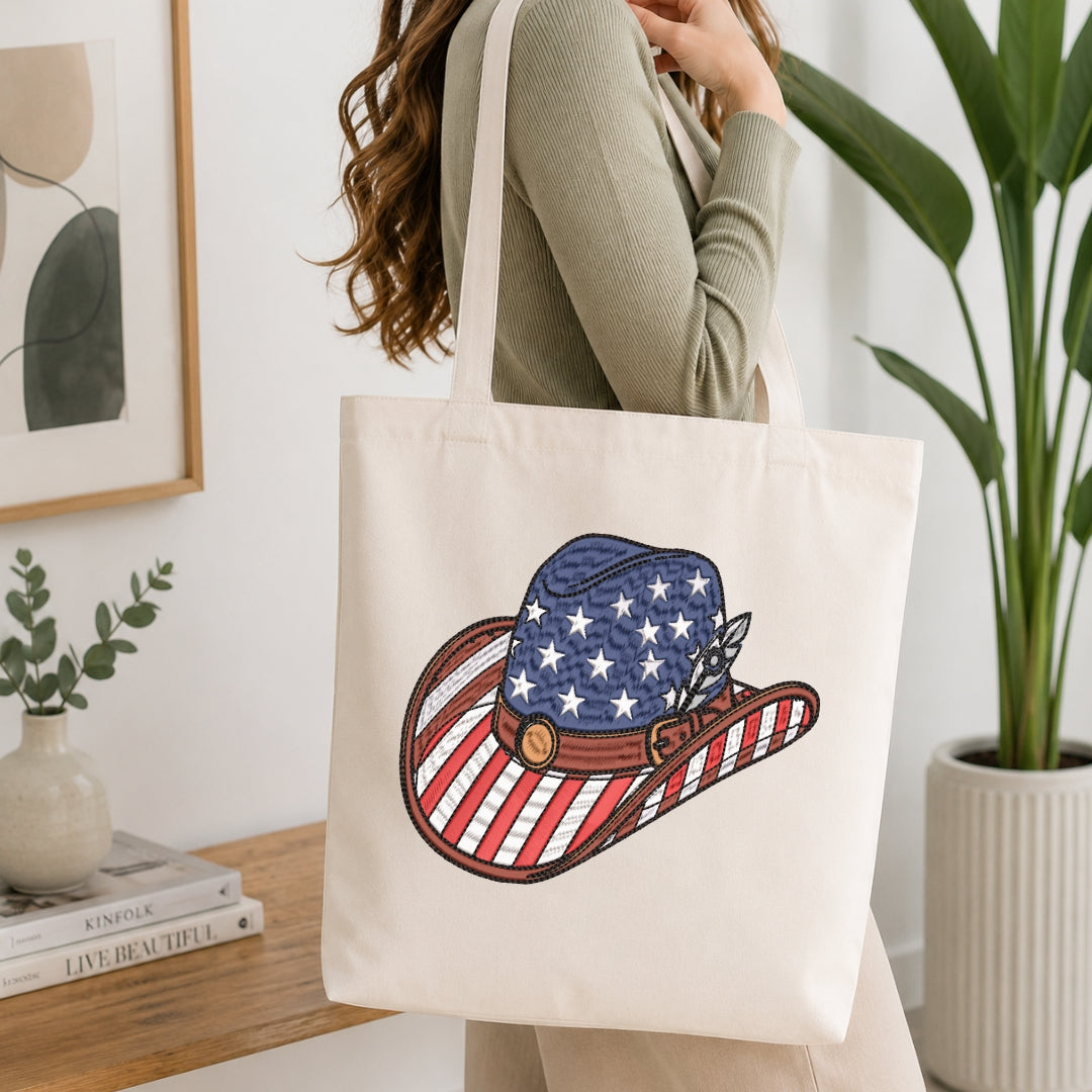 A person wearing a sage green ribbed long-sleeve top carries a cream canvas tote bag with a printed cowboy hat design featuring an American flag pattern. The scene is set in a bright, modern room with a wooden side table, a framed print, a small vase, and a potted plant.