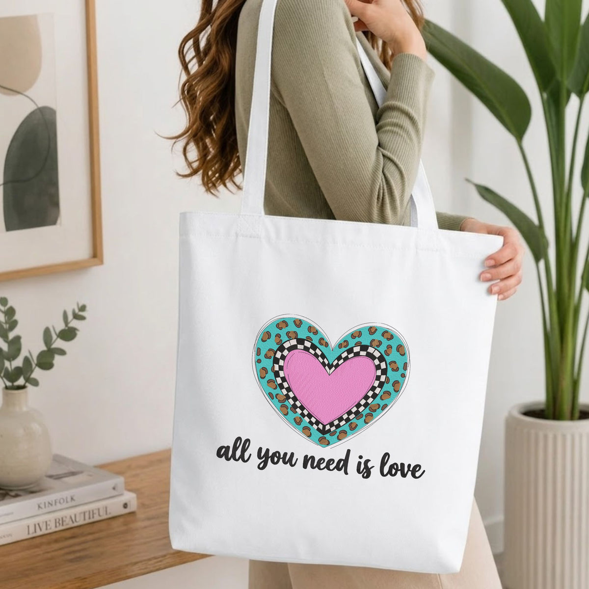 Woman carrying a white natural canvas tote bag featuring an "all you need is love" heart machine embroidery design in a bright, modern interior setting   